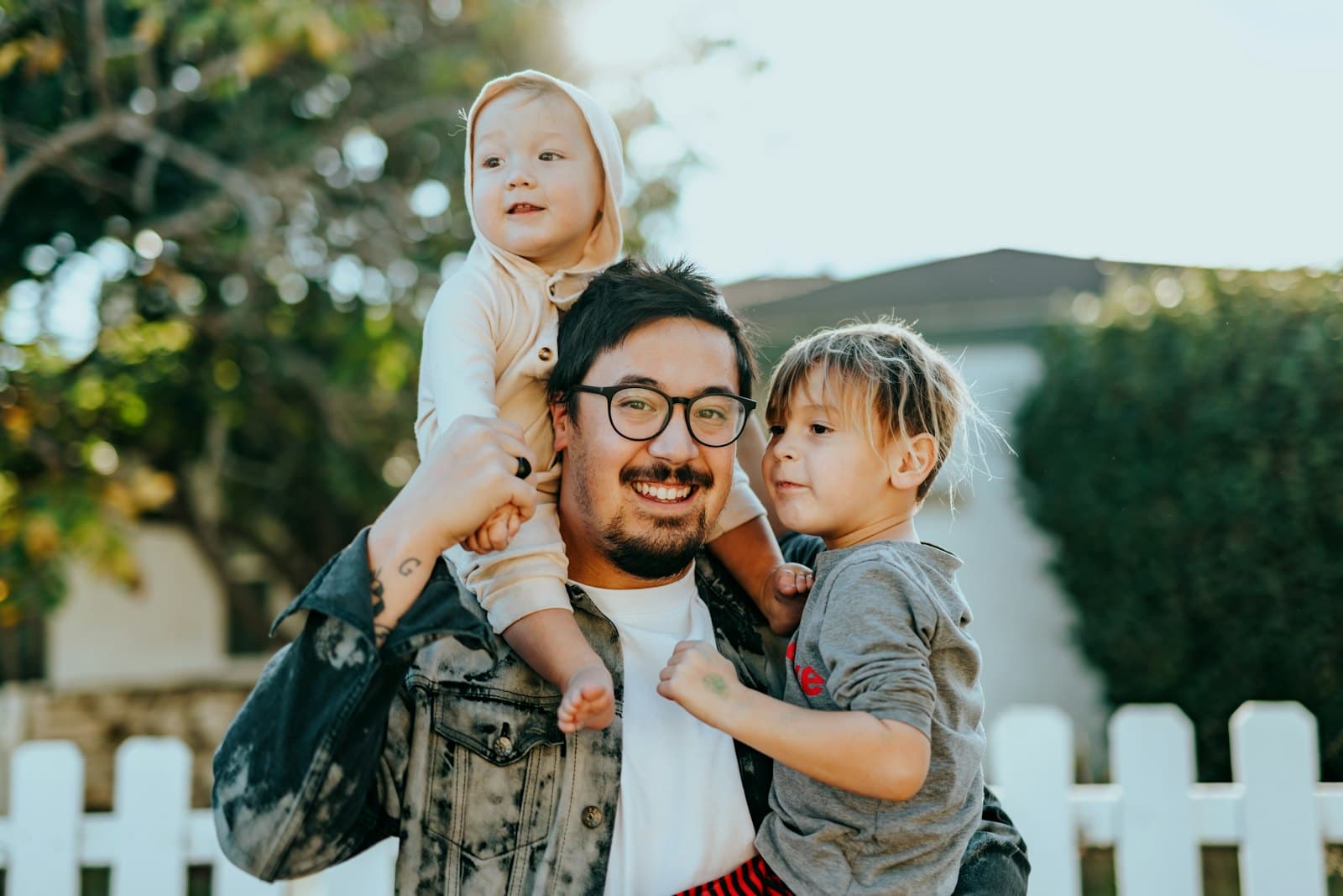 A father outside his home with his two young children on his shoulders